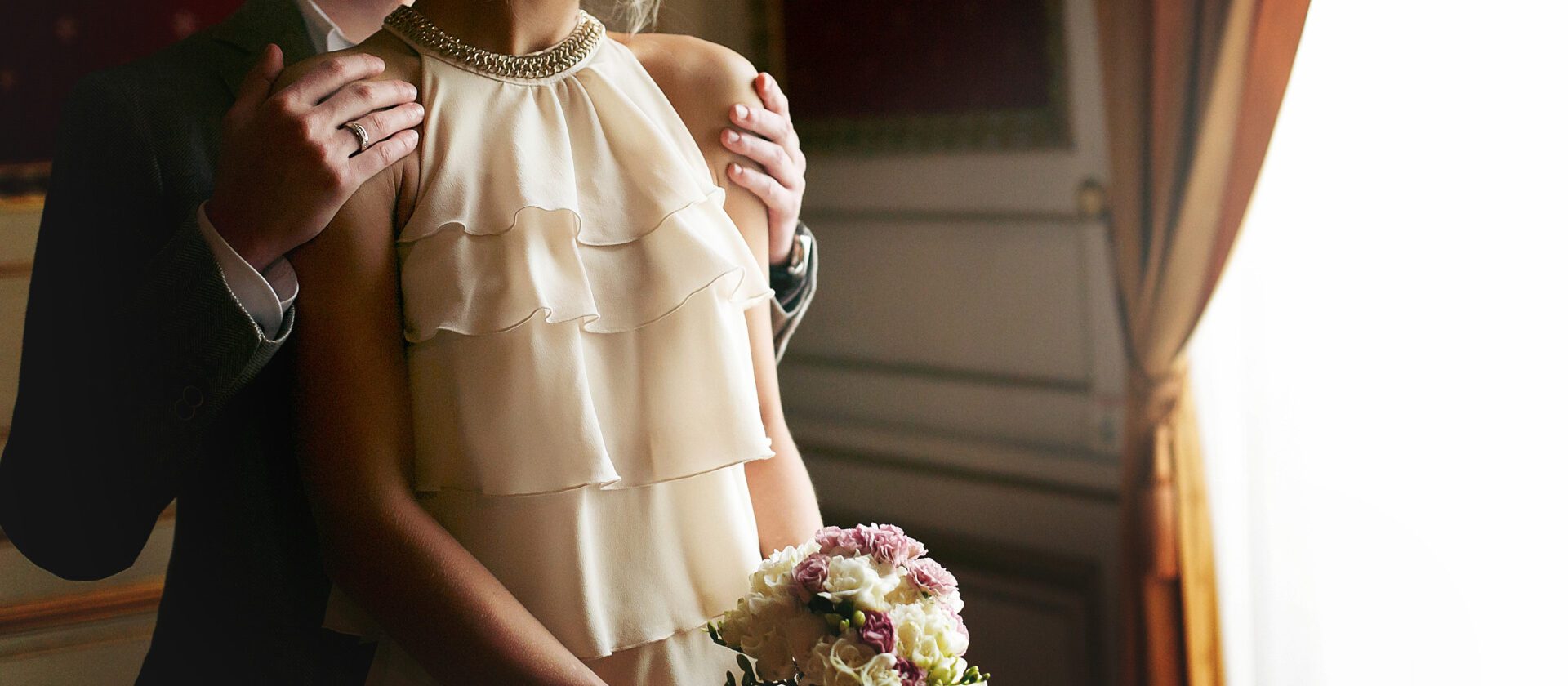 luxury bride in vintage dress with bouquet and stylish groom embracing and hugging at window in morning light in hotel room. happy wedding couple, sensual romantic moment